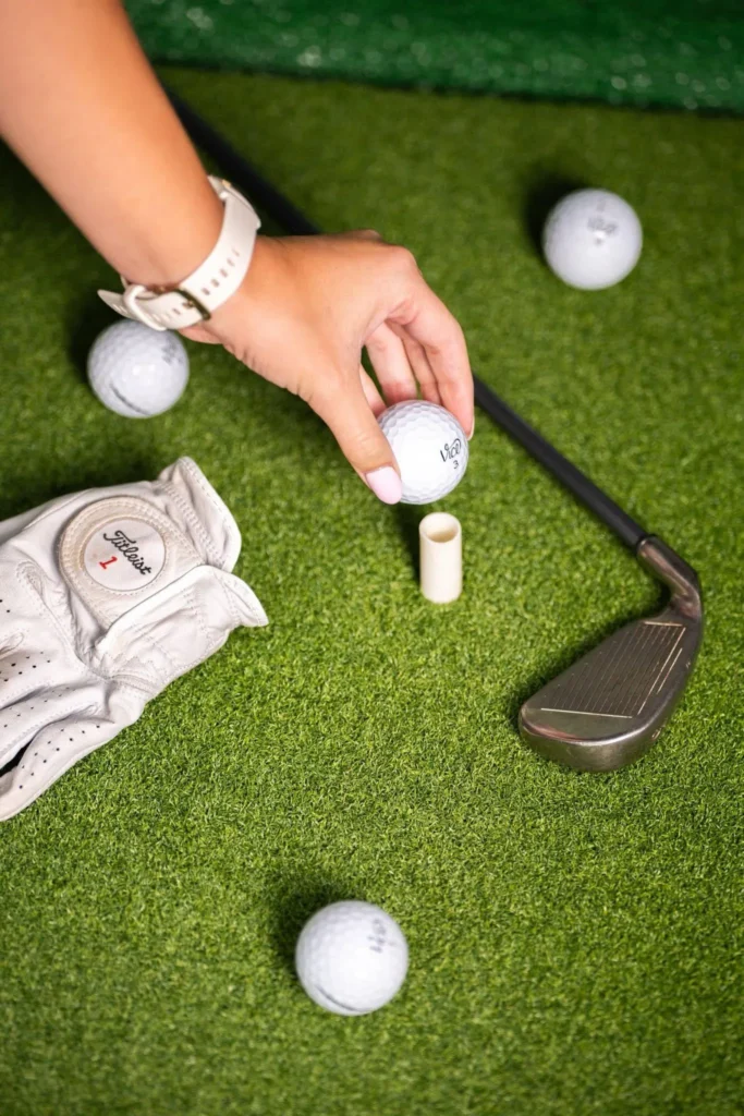 Hand placing a golf ball on a tee beside a wedge, glove and balls on simulator turf