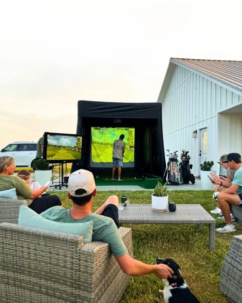 Guests relaxing around a Riverside Fairways mobile golf simulator in a backyard at dusk