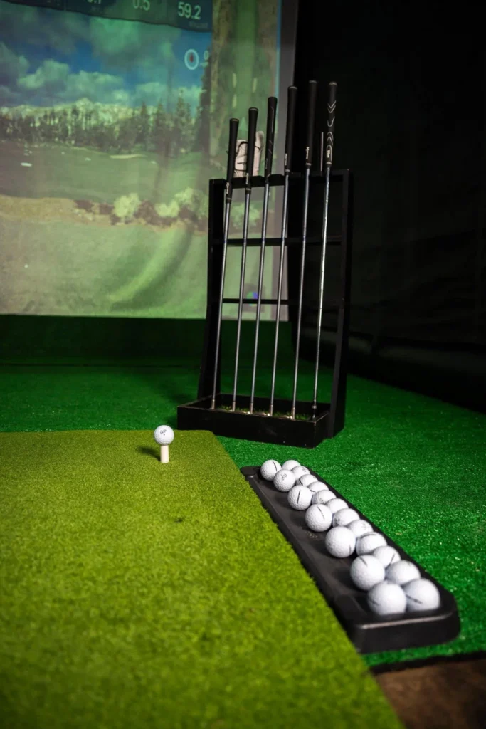 Mobile golf simulator set up indoors with a club rack and tray of balls on turf