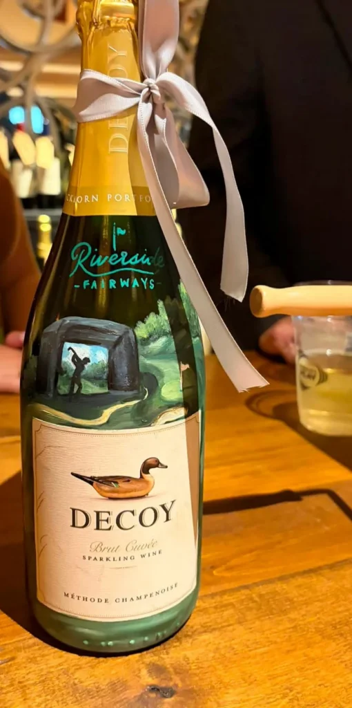 Sparkling wine bottle with a hand-painted Riverside Fairways golf simulator label, sponsored by Riverside Fairways for the wine pull at Fete Rouge 2026
